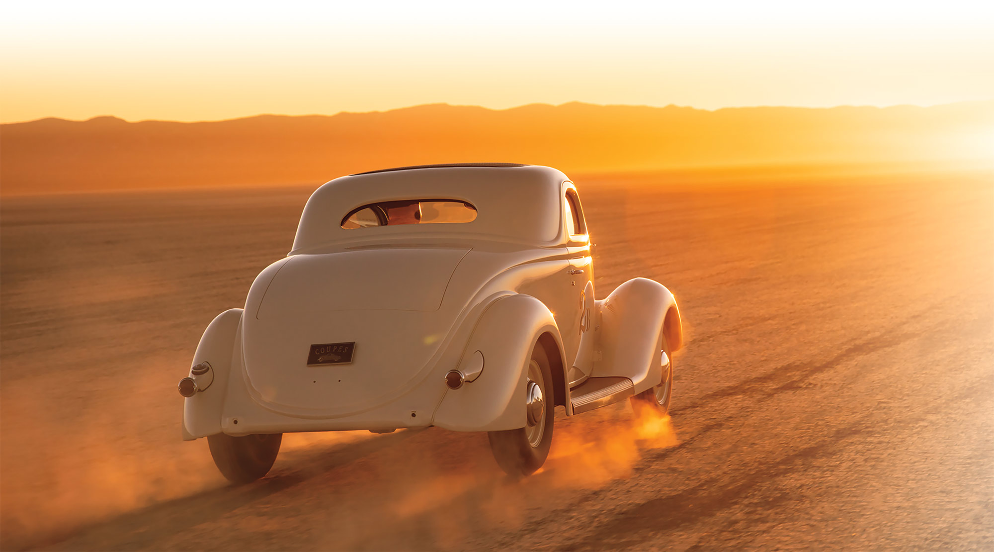 view of 1936 Coupe driving away on a dirt road during sunset