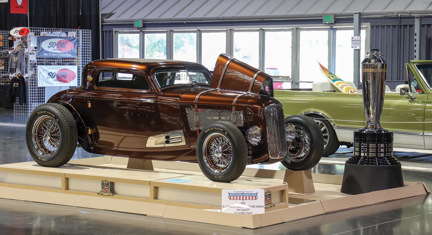 passenger side view of a metallic brown ’32 Ford three-window coupe on blocks with a large trophy standing beside