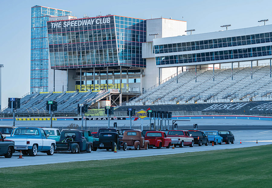 Rows of cars lined up in front of a big, square building with tons of windows and a sign that says "The Speedway Club"