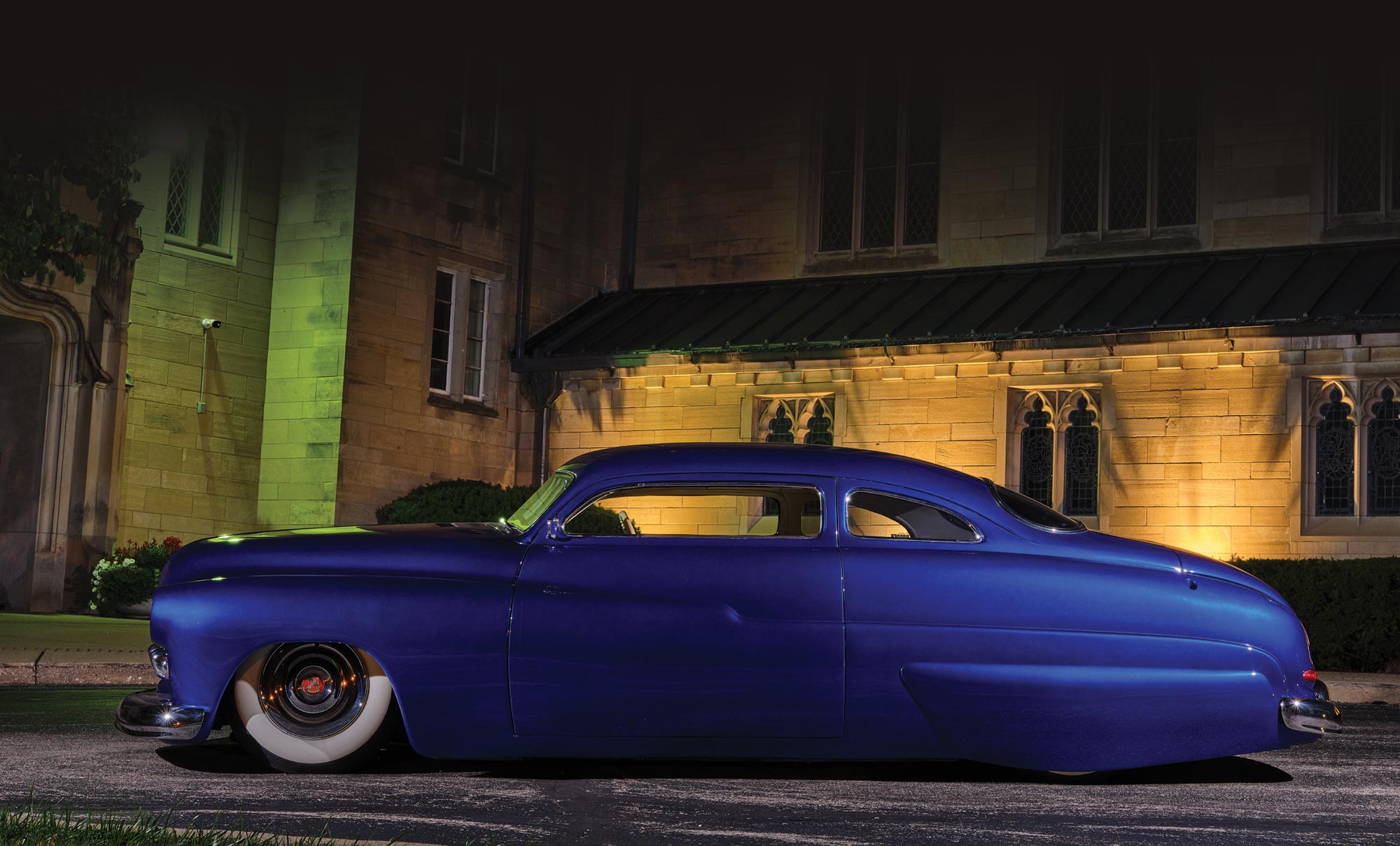 Driver's side profile view of the '50 Merc in Candy Cobalt blue, parked in front of a old stone building at night