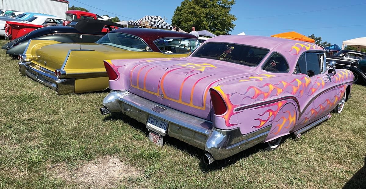 ’57 Buick with flames against a bright purple background parked next to a desaturated gold ’57 Cadillac