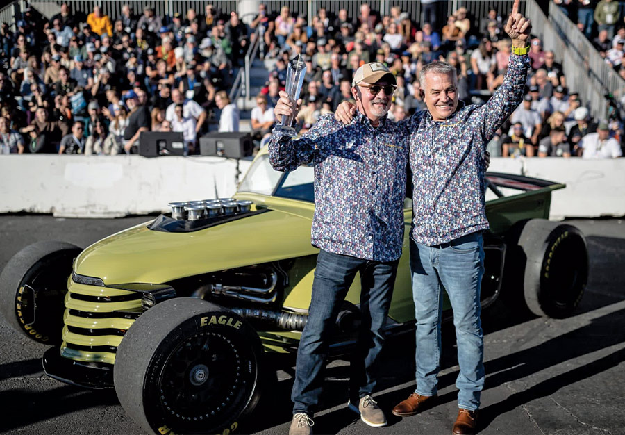 Two men in matching shirts posing triumphantly in front of their green hot rod and a crowd of people