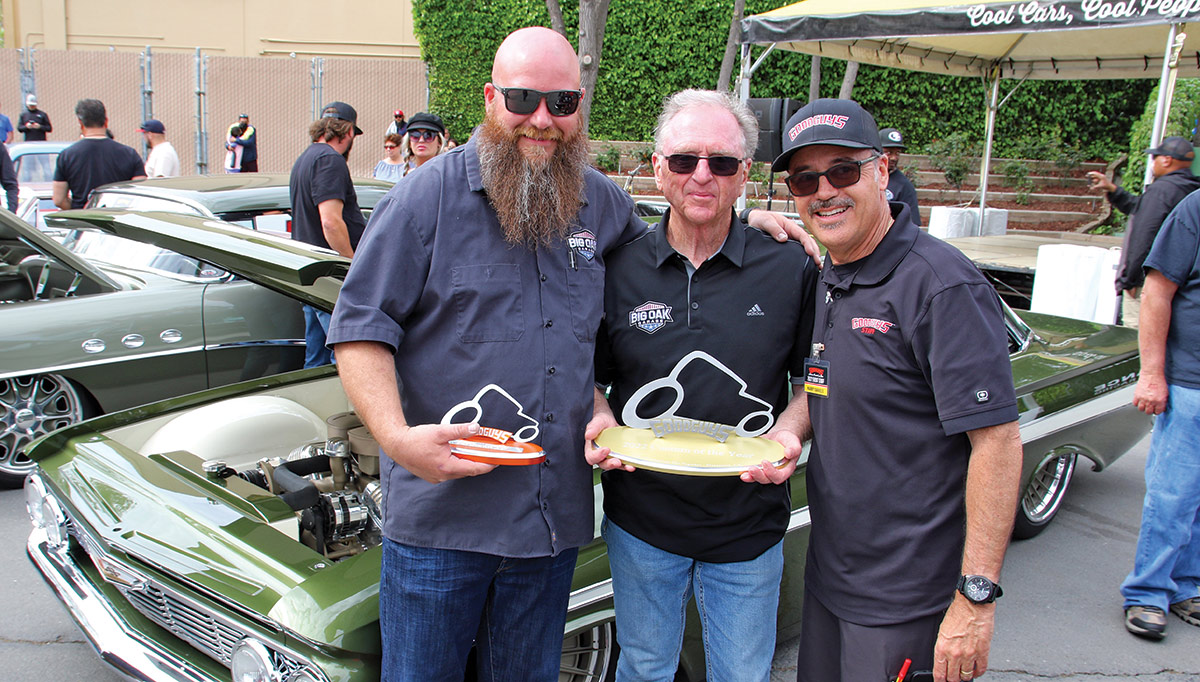 A group of three men in front of a show of cars, two of them holding trophies with cars on top