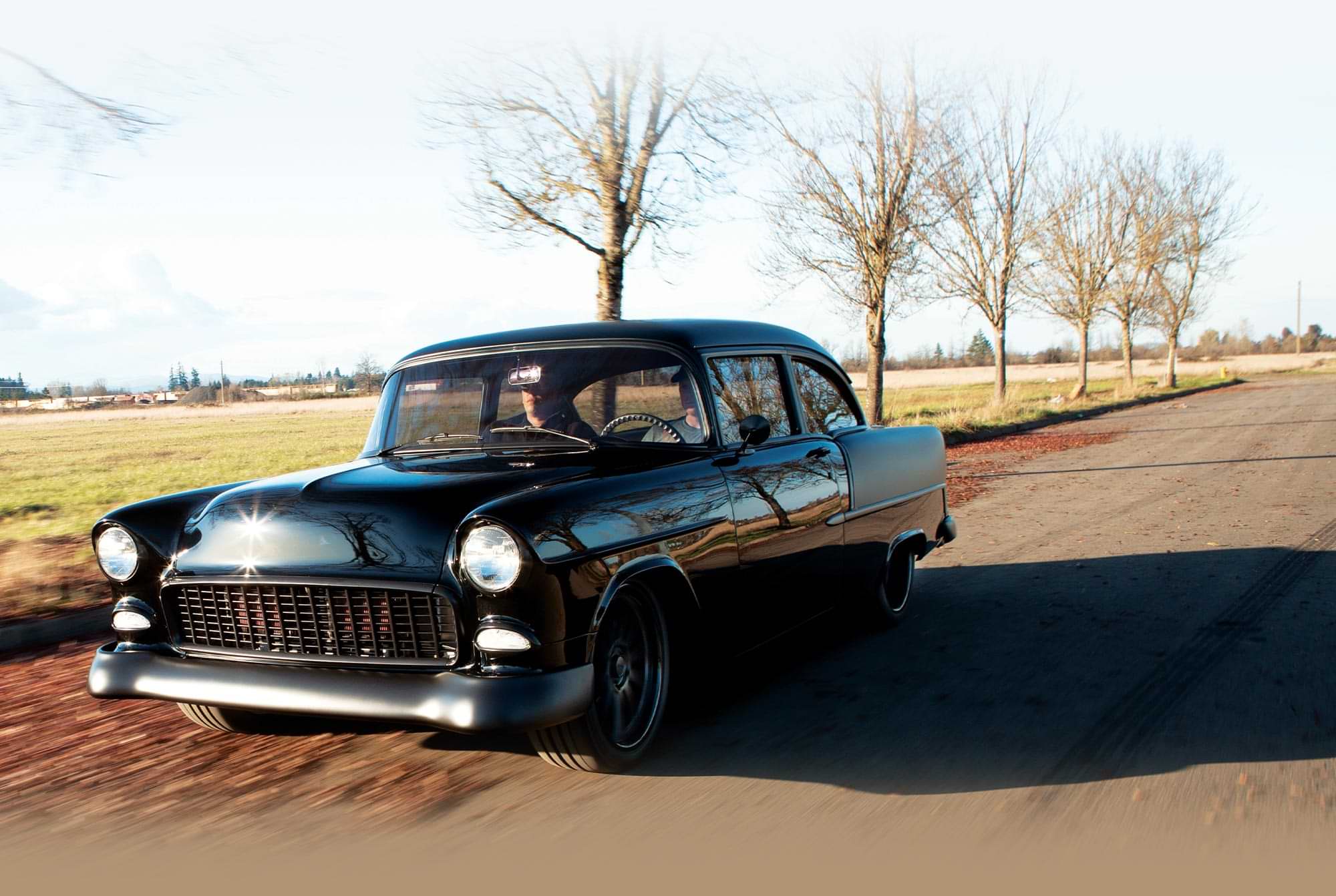 three quarter driver's side view of a black ’55 Chevy riding down a tree lined road