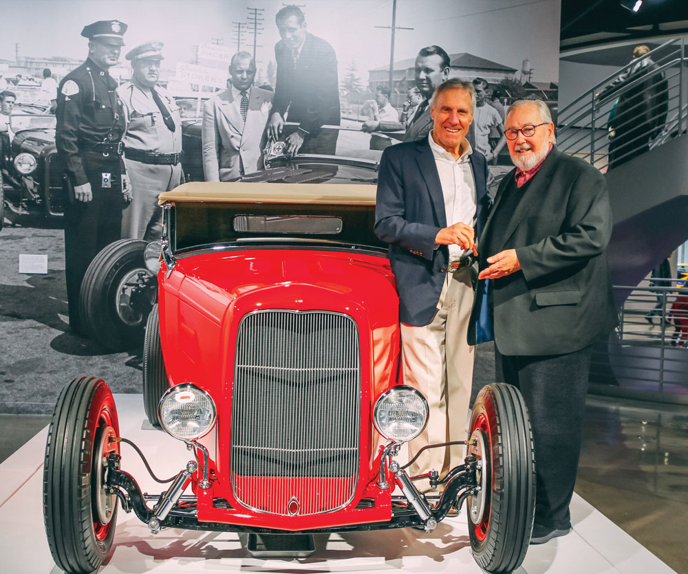 2 men standing next to red roadster