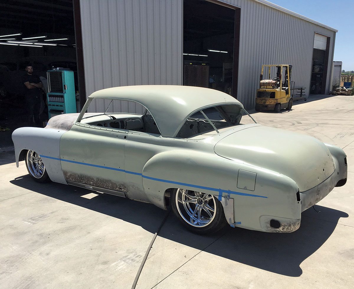 Rear side angle view of the original silhouette of the ’51 Chevrolet Bel Air car outside an auto shop area; The Chevrolet hardtop roofline carries a three-piece rear window with a unique C-pillar shape. The perfect stance comes from the Genesis chassis employing Chris Alston’s Chassisworks suspension components.