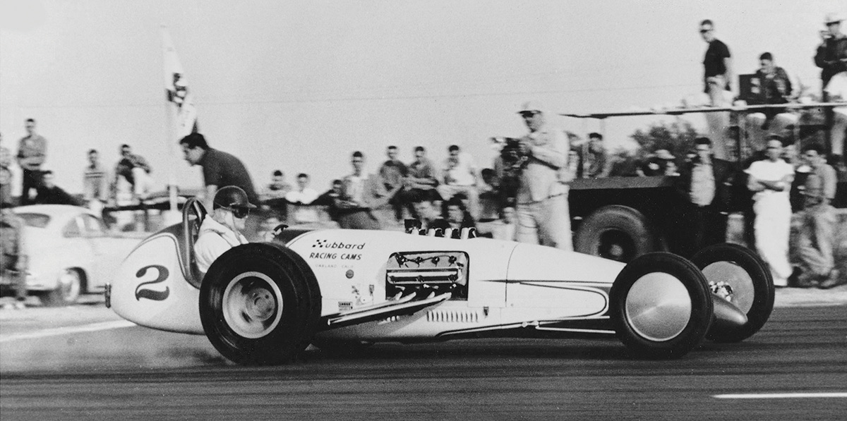 Black & white photograph of accentuating motion and body symmetry, Tommy’s spiked flame scallops imitated speed on Hubbard Racing Cams’ fuel dragster at Vaca Valley Raceway, 1958, with Dick Hubbard driving. The DeSoto-powered slingshot was built by Jack Friedland. The car had style and balance but did not perform to class standards.
