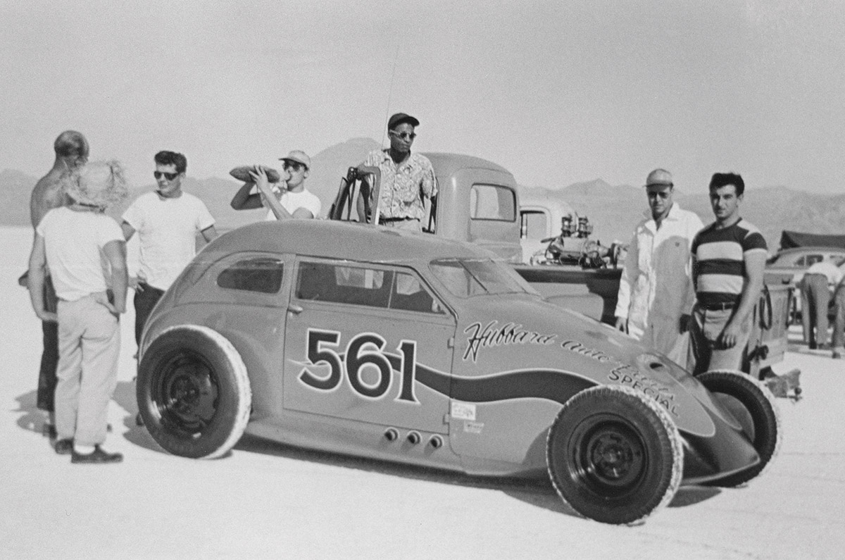 Black & white photograph of since the first Oakland Roadster Show, Tommy was close to the Hubbard racing family in Oakland. Here his work graces the nose and body of the Hubbard B/Comp flathead-powered Crosley at Bonneville, 1952. Running a de-stroked motor, the car set B/Comp sedan record at 131.96 and took a second in C/Comp with a 296-inch engine. Those identified include a young Romeo Palamides (’striped shirt) and driver Richard Hubbard (rear of car).
