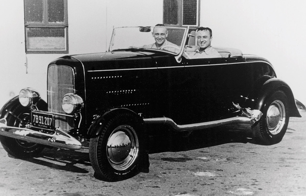 Black & white photograph of The Edelbrock-Bosio-Ladley-Edelbrock roadster. In 1956, the black ’32 bearing Tommy’s simple ’striping, won the AMBR title at the Oakland Roadster Show. Then-owner Eddie Bosio, San Francisco, is at the wheel and is joined by his sidekick, premier San Francisco engine builder Charlie Tabucchi. Bosio bought the roadster in the early ’50s from speed parts wizard Vic Edelbrock Sr. and transformed the spare and fast dry lakes racer into this street cruiser.
