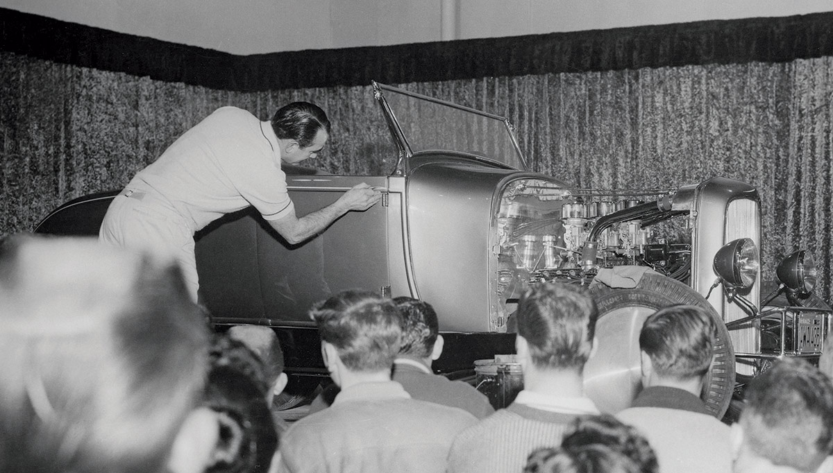 Black & white photograph of applying the final accent line on Ken Fuhrman’s A-V8, which was a display buildout in the Romeo Palamides Automotive booth. Oakland Roadster Show, 1958.