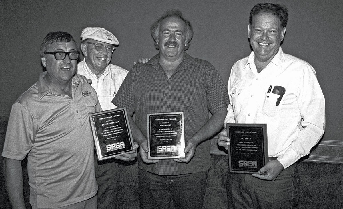 Black and white photograph of (from left to right) Gray Baskerville, Tom Medley, Andy Brizio, and Tex Smith holding Street Rod Hall of Fame award plaques as they smile posing for a picture together
