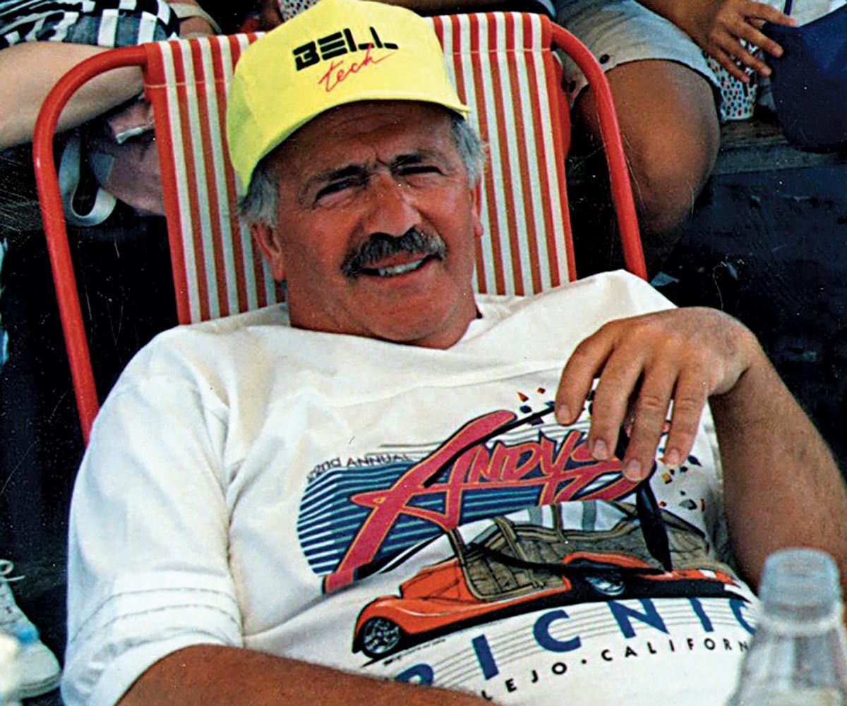 Close-up portrait vintage photograph perspective of Andy Brizio (known as The RodFather) smiling as he is laid back sitting down on a red/white striped lawn chair while wearing a yellow Bell Tech hat and white graphic t-shirt