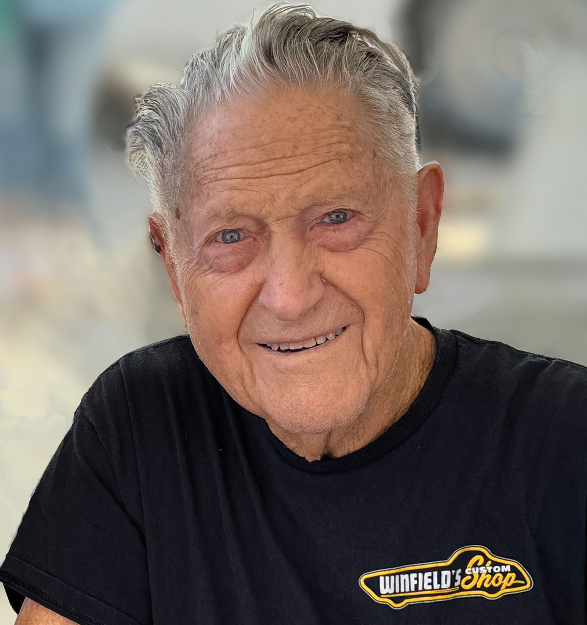 Close-up portrait headshot photograph perspective of Gene Winfield smiling in a black graphic t-shirt that shows a small Winfield's Custom Shop white/yellow logo