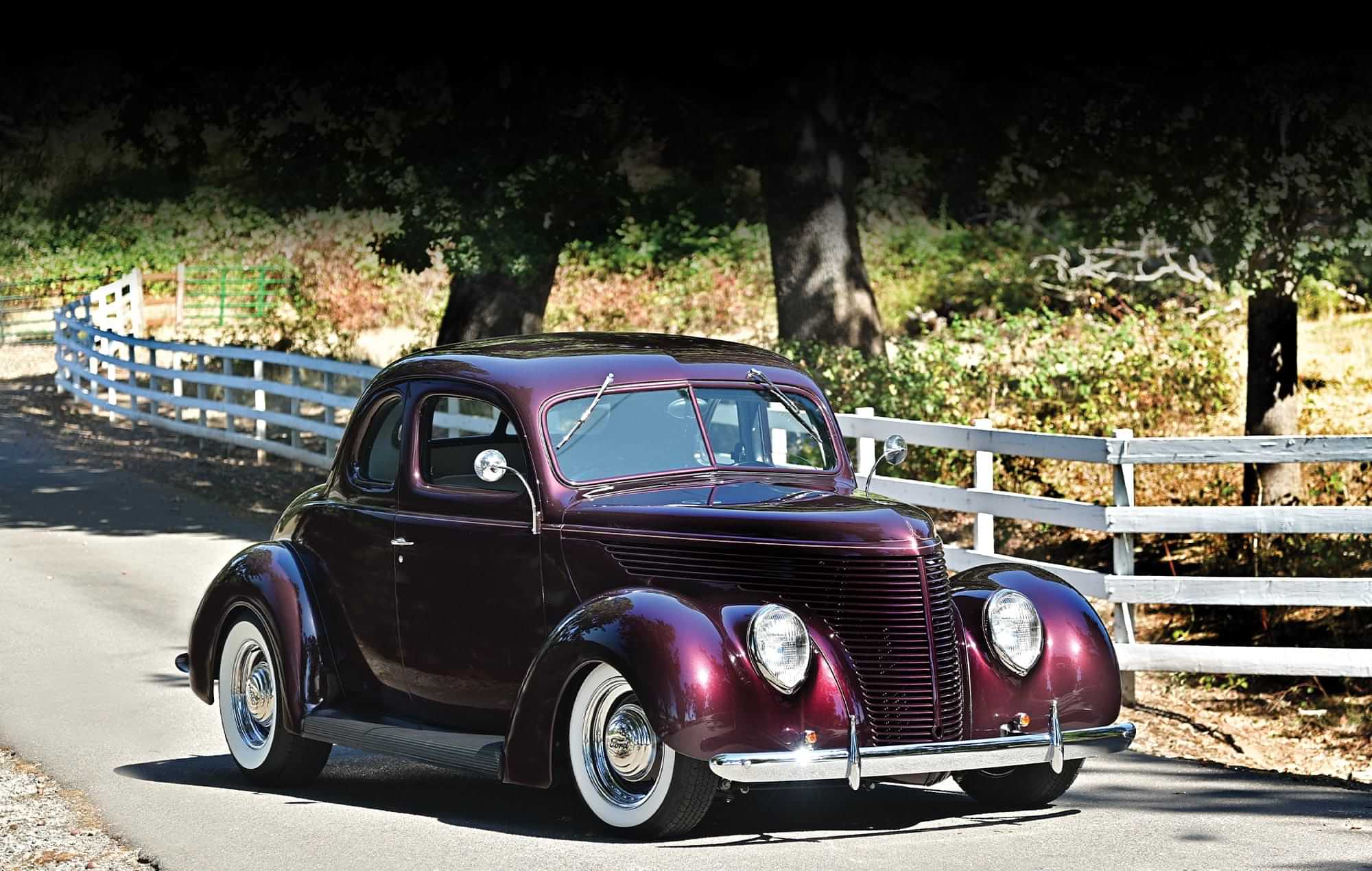 front 3/4ths view passenger side view of the deep burgundy ’38 Ford Standard Coupe parked on a narrow road beside a white picket fence and large trees