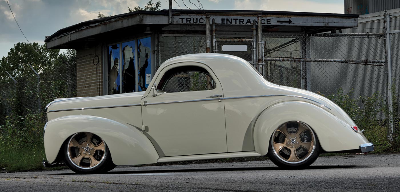 drivers side profile of the desert beige ’41 Willys Coupe parked next to a fenced off dilapidated factory