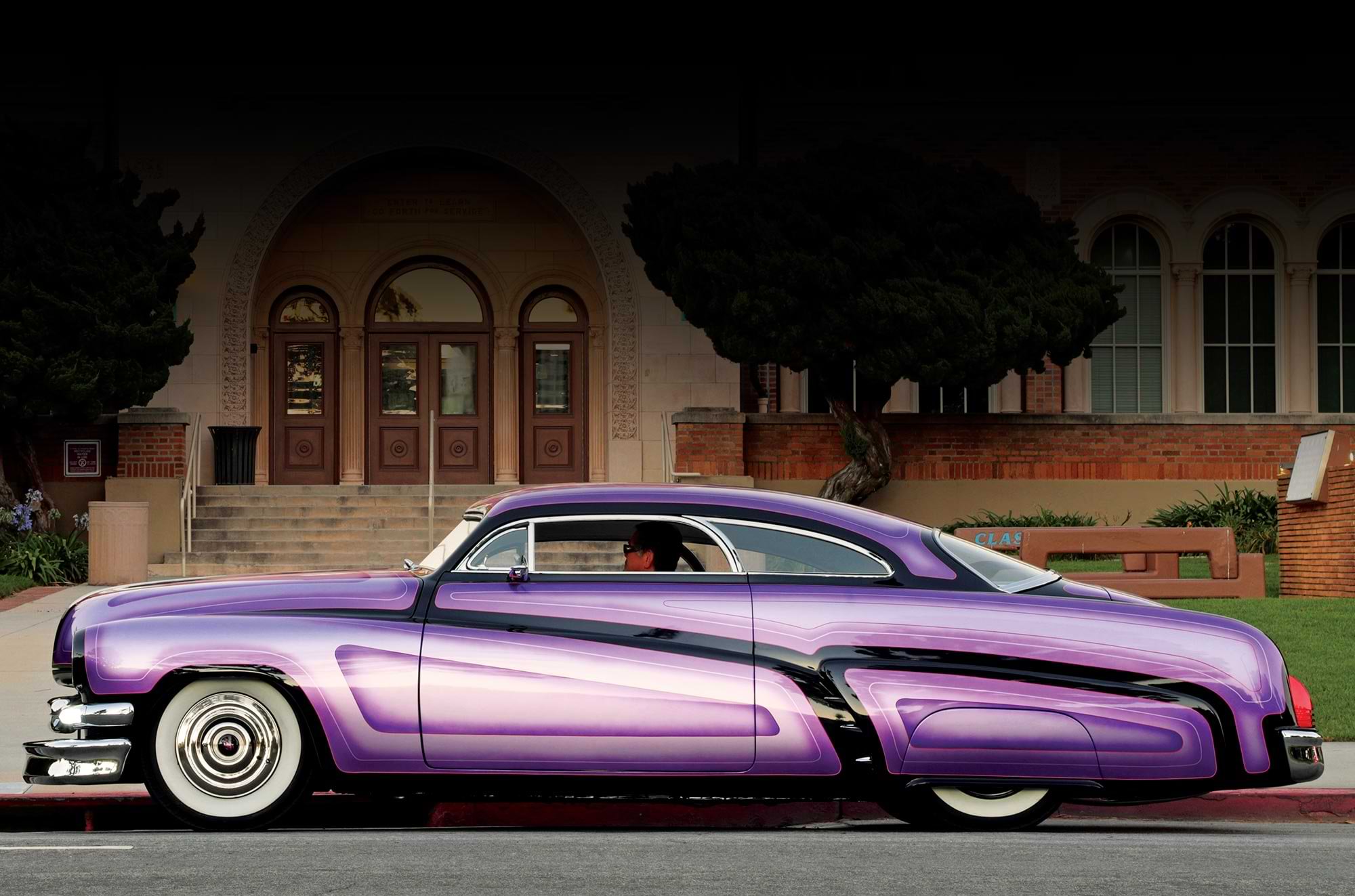 driver side profile of the custom vibrant purple ’51 Mercury parked in front of a dated red brick building