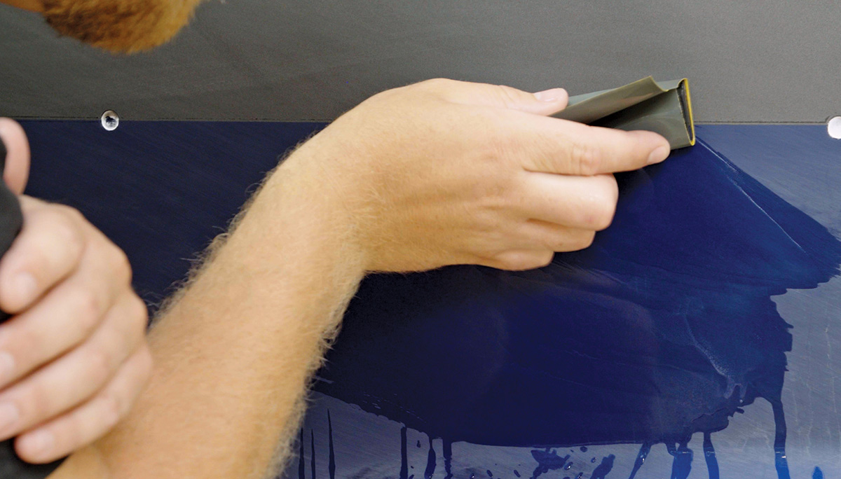 Close-up landscape photograph perspective of a person's hand applying a 600-grit grey sand cloth object onto the painted surface of a vintage vehicle