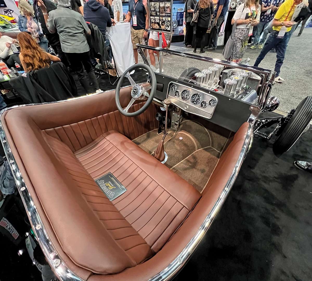 Portrait interior photograph perspective of the 1923 Isky tribute Ford roadster car seat on display at the SEMA show as there are other spectators nearby; The tribute roadster features Stewart-Warner gauges with a reproduction Auburn panel featuring engine turning. The replica banjo wheel is from Grant. The tach is column mounted on the original roadster and dash mounted on tribute roadster.