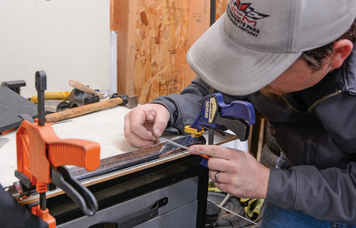 a mechanic leans in close to file an area on a piece of trim clamped to a work table
