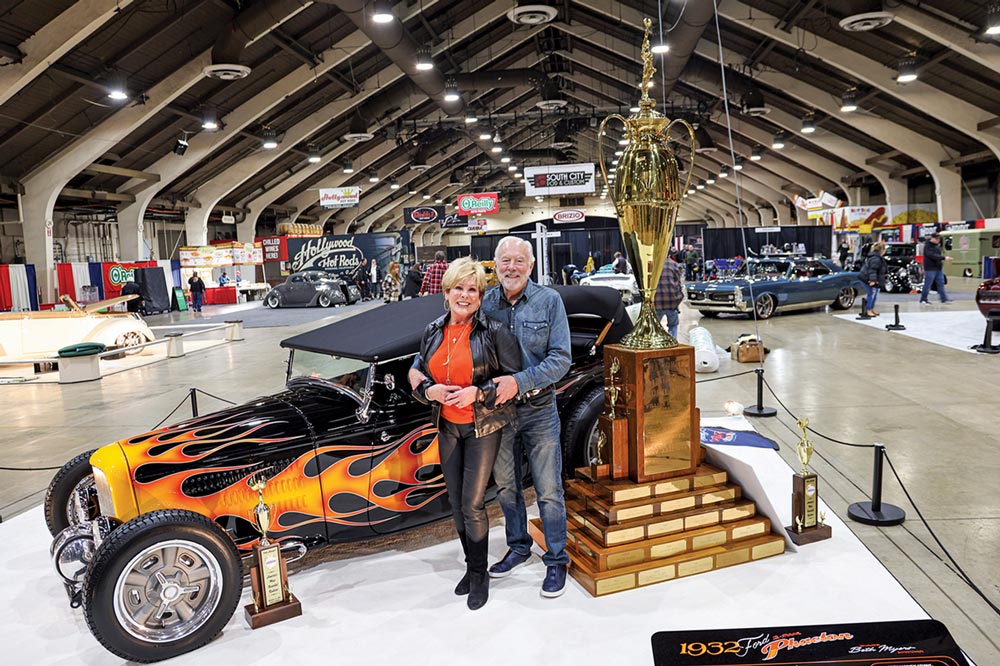 Beth and Ross Myers stand together in front of their black ’32 Ford phaeton with flames with a tall trophy standing beside them