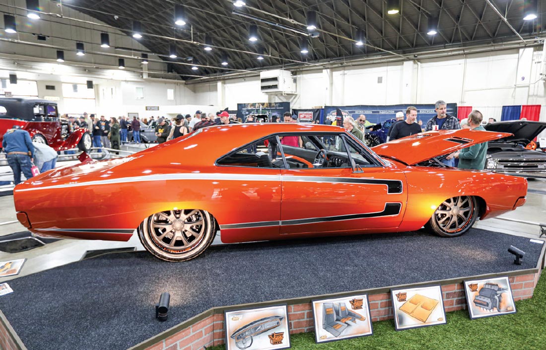 passenger side view of a vibrant orange ’68 Dodge Charger with its hood lifted stationed on a carpeted mock brick wall display