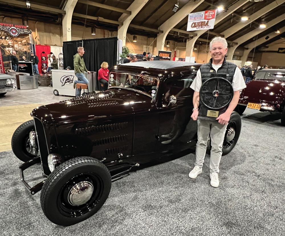 a man holding a steering wheel shaped award smiles while standing beside a dark brown ’32 Ford highboy five-window coupe