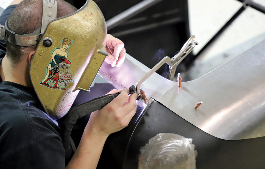 person wearing a welding helmet adding a metal strip to the panel above the rear window