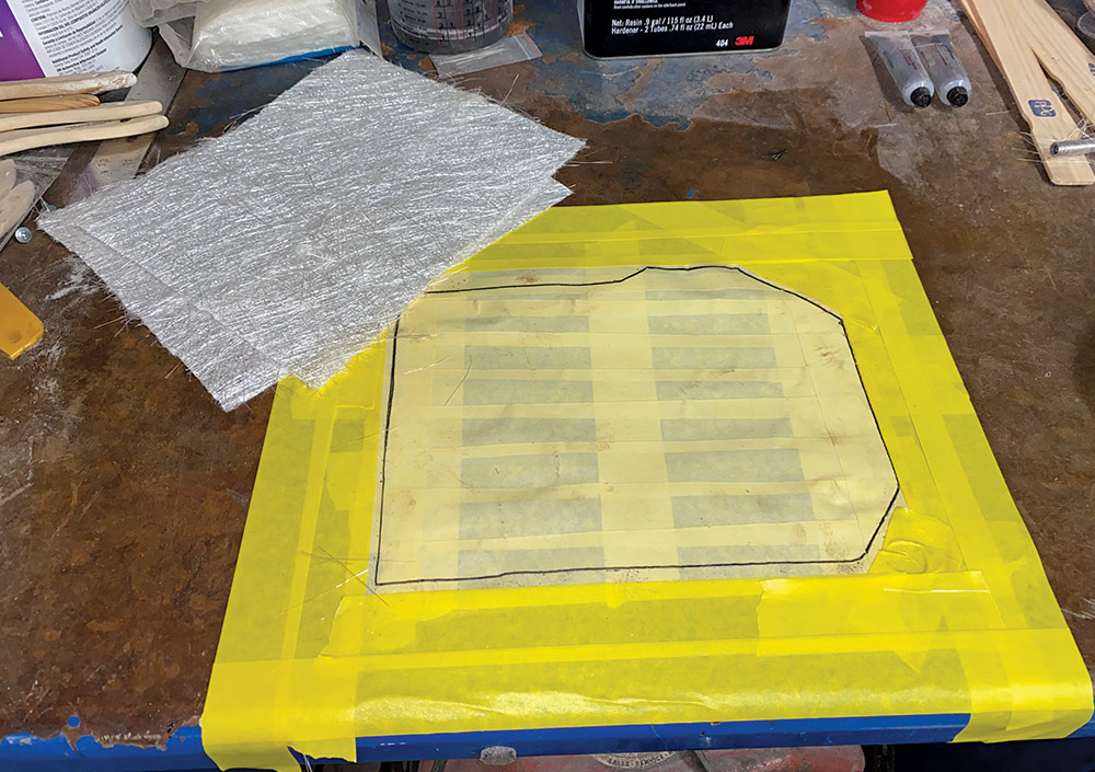 masking tape with engine compartment hole traced on it on workbench next to fiberglass mat
