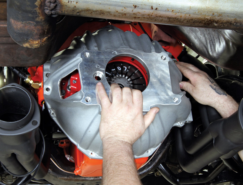 Hands adjusting a bellhousing on a red engine block under a vehicle.