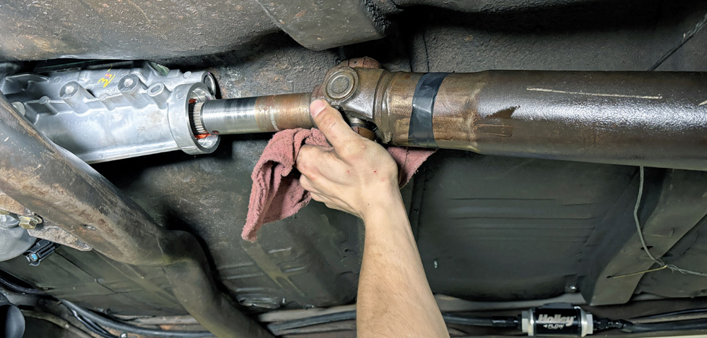 A hand holding a pink cloth is working on a drive shaft beneath a car.