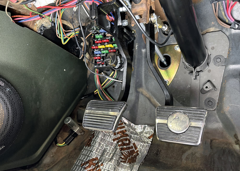 Interior of a vehicle showing clutch and brake pedals, a fuse box with multicolored wires, and a speaker in the dash.