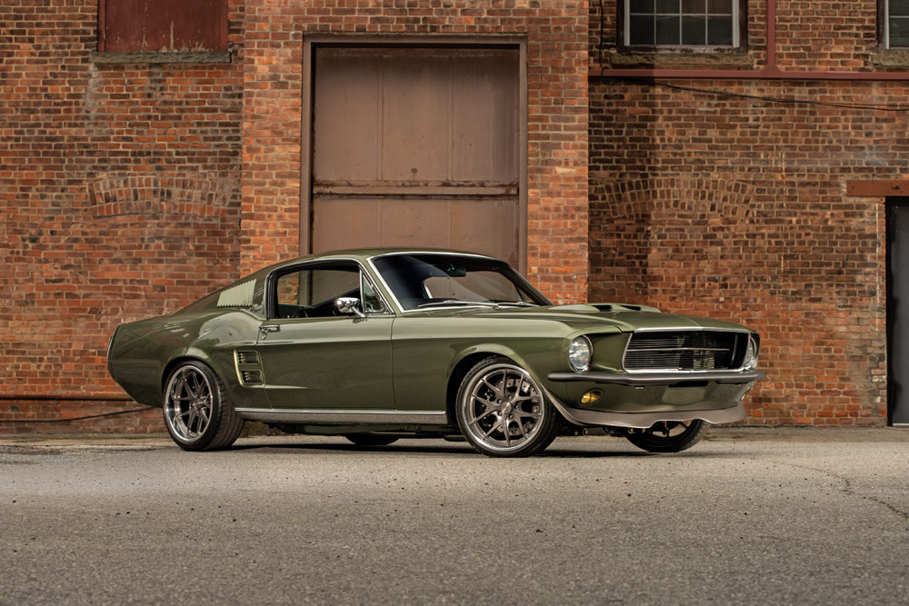 A classic olive green Ford Mustang parked in front of a brick building.