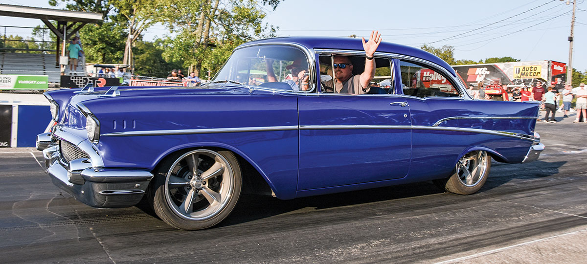 a driver waves while riding in the 2024 Golden Star Giveaway Car, a vibrant navy blue 1957 Chevy two-door sedan