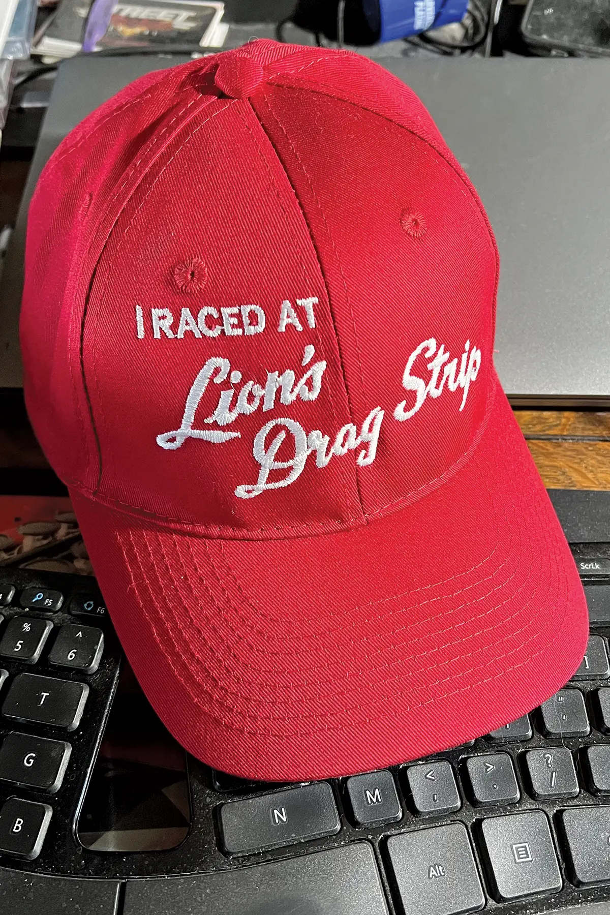 Portrait orientation close-up photo of a red hat that states I RACED AT Lion's Drag Strip, in which the hat is situated on top of a computer keyboard