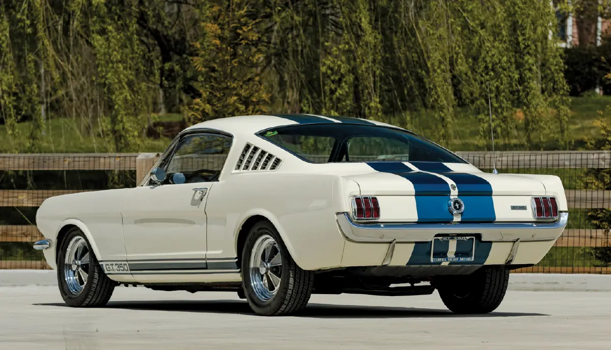 Landscape orientation outdoor close-up photograph rear three-quarter view of the 1965 Ford Mustang Shelby G.T. 350 in Wimbledon White adorned with Guardsman Blue Le Mans stripes parked nearby a wooden fence with greenery and trees in front in the far distant background