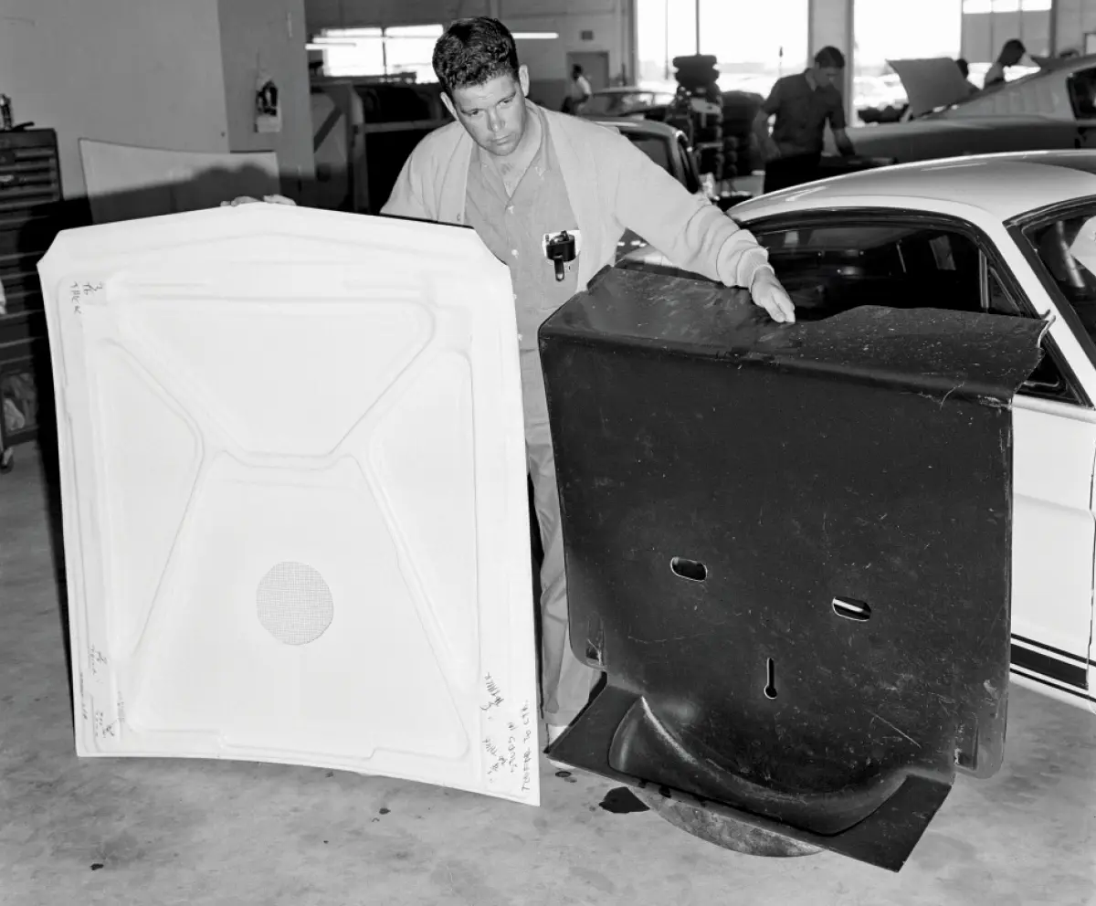 Portrait orientation vintage black and white photograph view of a man holding a fiberglass hood (left) and the rear interior floor piece (right) that replaces the factory rear seating area and now serves as the new location for mounting the spare tire