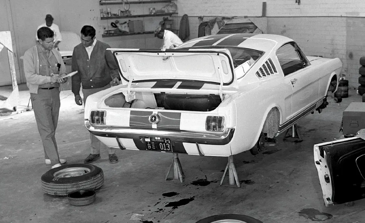 Landscape orientation vintage black and white photograph view of men inside a paint shop examining a crucial vital document sheet for the Shelby Mustang is now outfitted with the upgraded suspension and then readied to go out the door; This shot was taken at the Shelby facility at LAX.