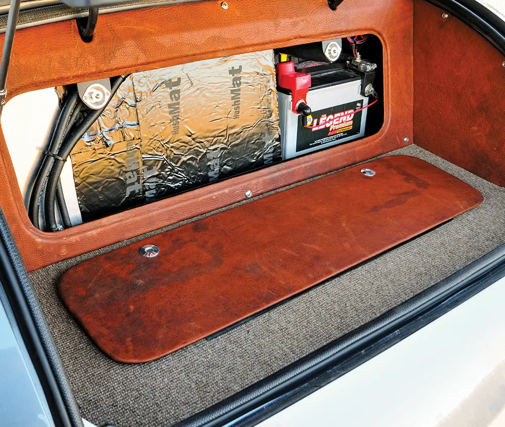 Trunk interior with a brown panel on a gray carpet and a mounted battery.