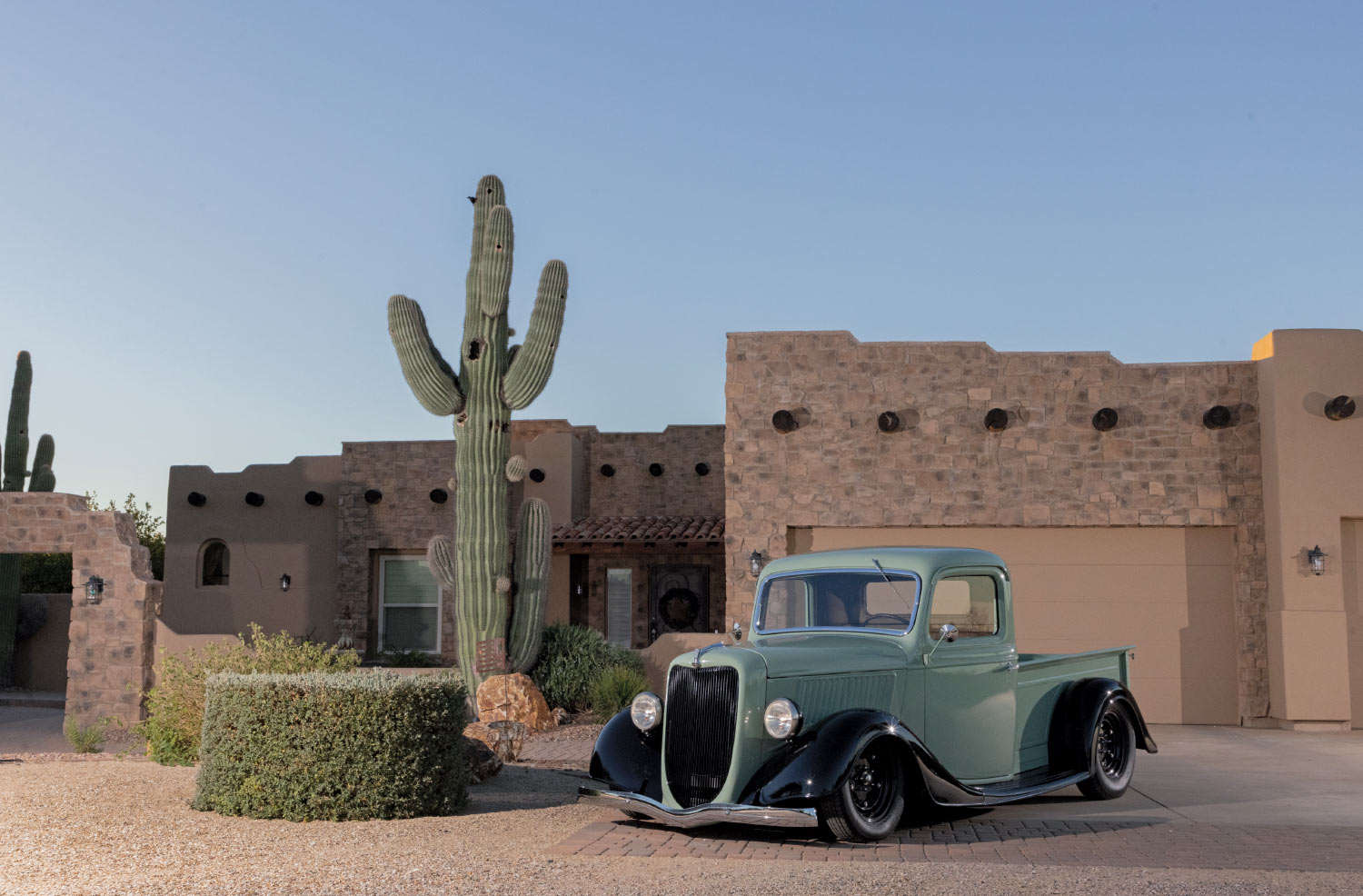 sage green 1936 ford pickup truck parked in front of brick house next to tall cactus in desert setting