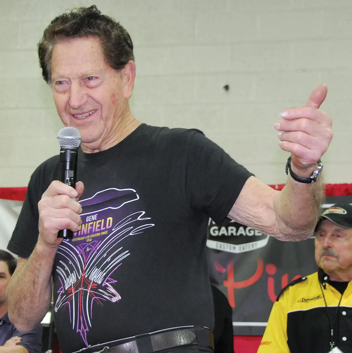 Portrait orientation photograph close-up view of Gene Winfield smiling as he is standing wearing a black t-shirt containing graphic artwork on it while he is holding a microphone nearby his mouth with his left arm hanging in the air giving a thumbs up gesture while there are two men glancing around behind his back; Gene gave a talk at the 2008 Detroit Autorama, the year he was honored as the Builder of the Year