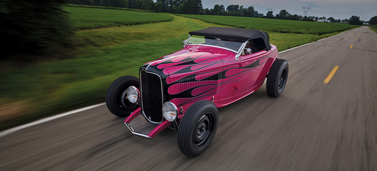 1932 Ford Highboy Roadster with black soft top and magenta flame job, cruising a scenic rural highway
