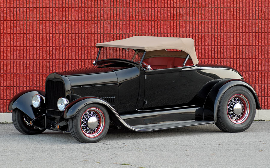 1930 Ford Roadster with tan top, red accents, and wire wheels against a textured red brick backdrop