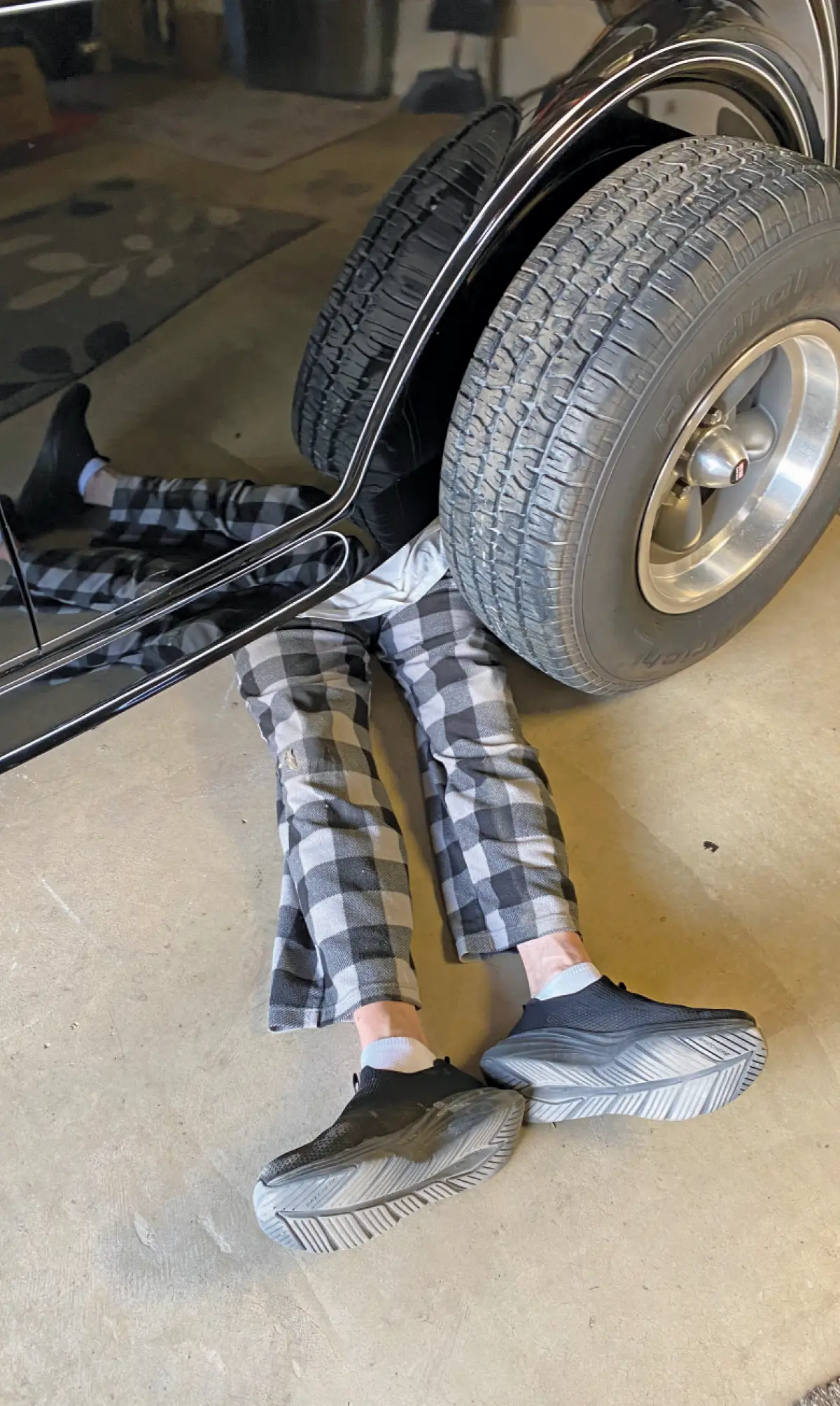 Portrait orientation indoor photograph of a person's legs, wearing gray and white patterned plaid sweatpants and dark black sneakers, are visibly seen under near the left passenger rear large tire side of the vintage hot rod 1932 Ford McMullen Roadster in black; They appear to be working on the vehicle inside a home garage setting