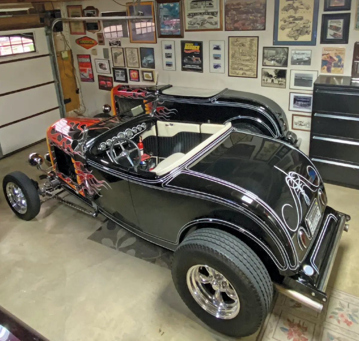 Landscape orientation indoor photograph side angle view of the vintage hot rod 1932 Ford McMullen Roadster and its half-roadster counterpart in black with yellow and orange flame decals are parked side-by-side in a home garage with numerous car themed framed pictures and posters on the walls with the home garage door closed