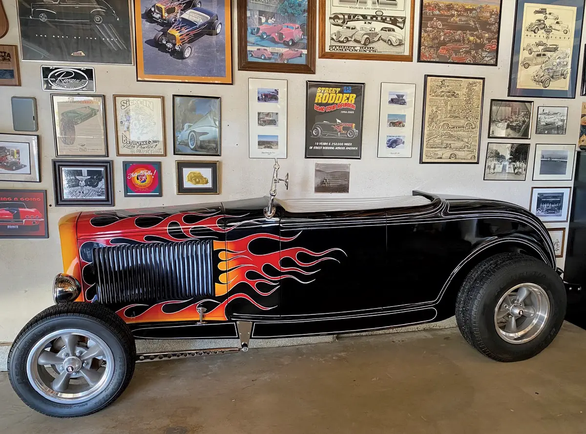Landscape orientation indoor photograph side angle view of the vintage hot rod 1932 Ford McMullen Roadster half-roadster counterpart in black with yellow and orange flame decals parked next to a home garage wall with numerous car themed framed pictures and posters on the walls