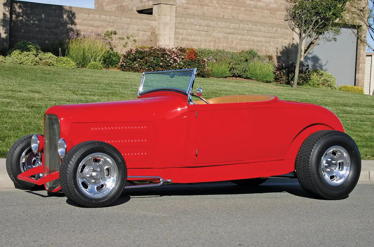 Red vintage hot rod roadster with chrome wheels and a tan interior, parked on a street in front of a landscaped wall.