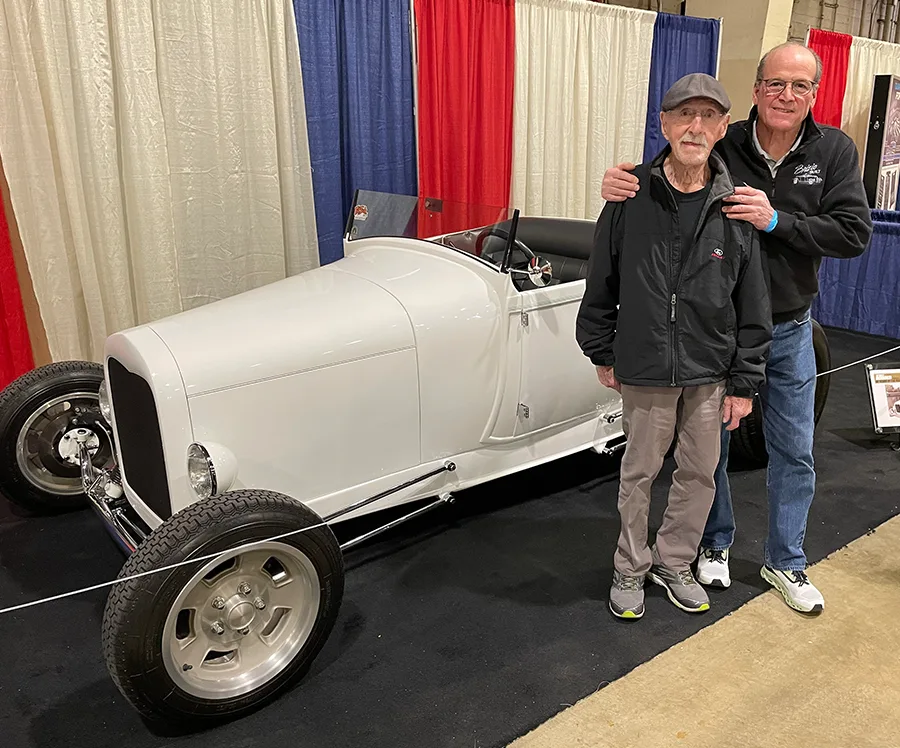 An older man in a cap and a younger man smiling and standing next to a sleek, white vintage-style hot rod roadster at an indoor car show with red, white, and blue drapes.