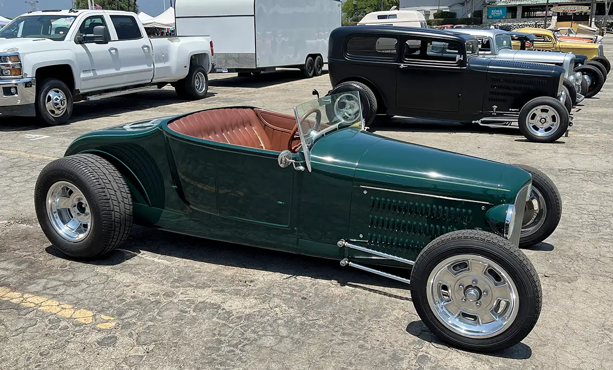 A custom dark green Ford track roadster hot rod with a tan interior and Halibrand wheels, parked outdoors with other classic cars in the background.