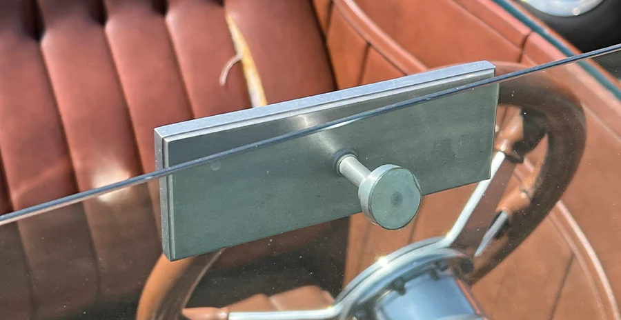 Detailed view of a classic hot rod's interior, showing a simple, streamlined aluminum rearview mirror attached to the windshield with a single knob, with the wood steering wheel and brown leather interior in the background.