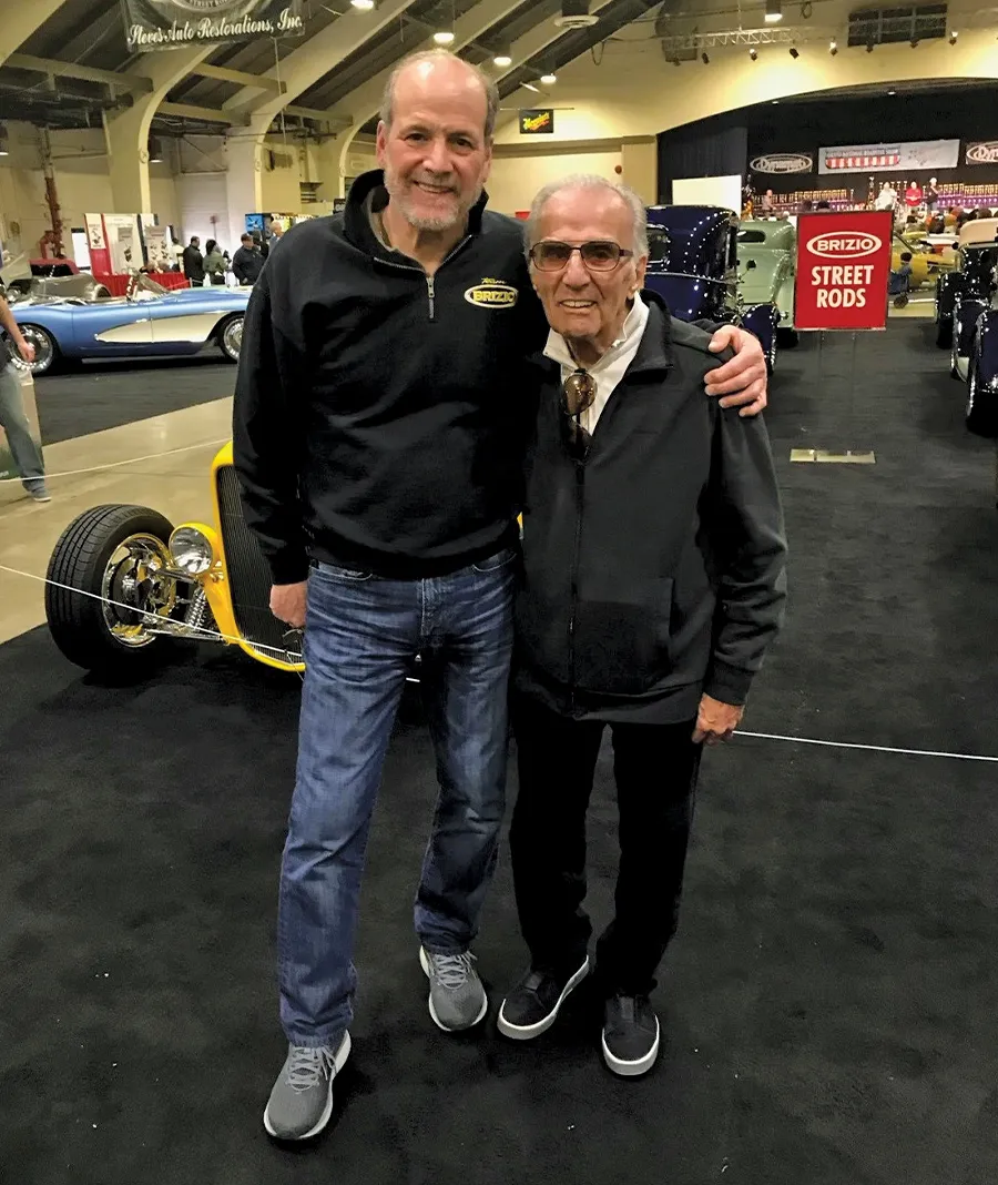 Brizio posing with Angelo at the Grand National Roadster Show with various street rods on display in the background